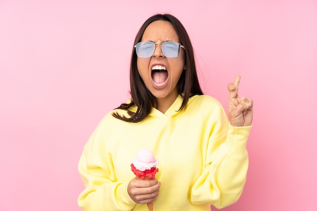 Young brunette woman holding a cornet ice cream on isolated pink with fingers crossing