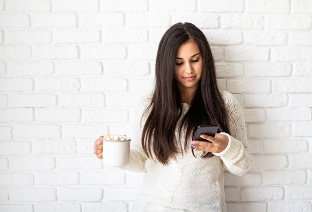 Giovane donna castana che tiene una tazza di cacao marshmallow e utilizzando il suo telefono cellulare