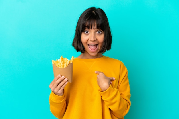 驚きの表情で孤立した青い背景の上にフライドポテトを保持している若いブルネットの女性