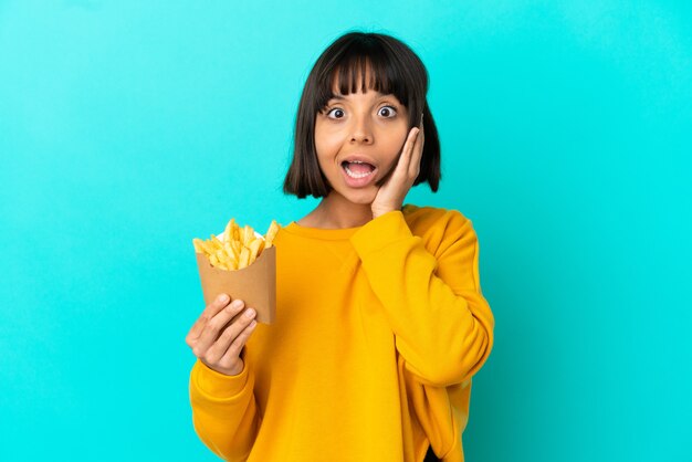 Young brunette woman holding fried chips over isolated blue background with surprise and shocked facial expression