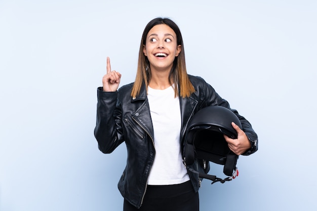 Giovane donna castana che tiene un casco del motociclo sopra la parete blu che alza un dito su