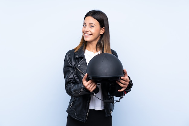 Молодая брюнетка женщина держит мотоциклетный шлем над синей стеной с счастливым выражением