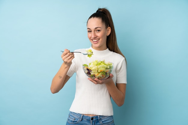 Young brunette woman holding a salad over isolated blue wall