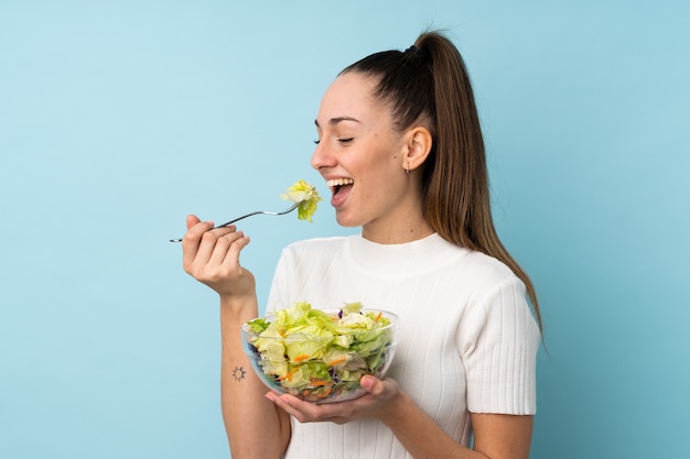 Young brunette woman holding a salad over isolated blue wall