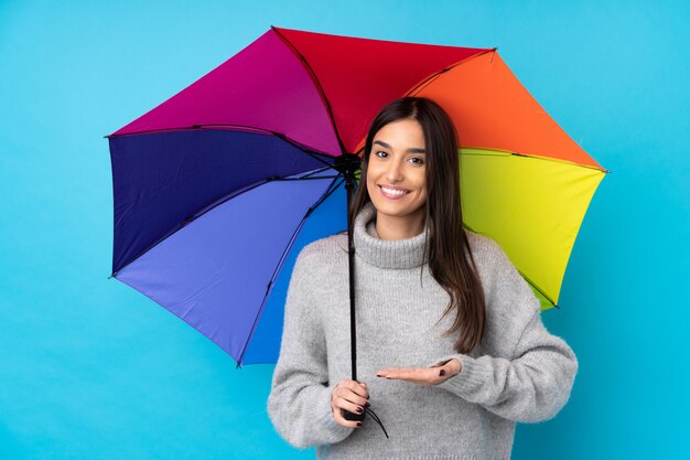 Young brunette woman holding an umbrella over blue wall extending hands to the side for inviting to come