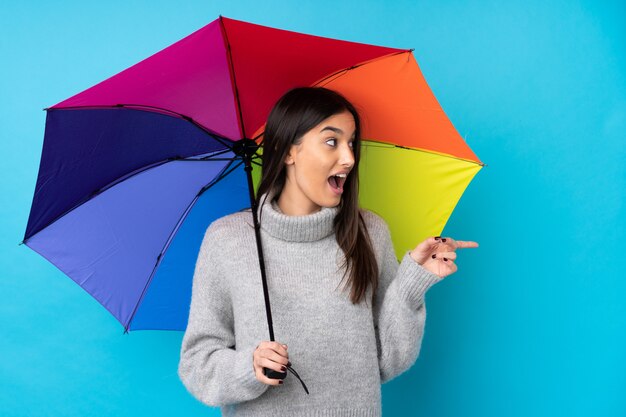 Young brunette woman holding an umbrella over blue wall pointing finger to the side