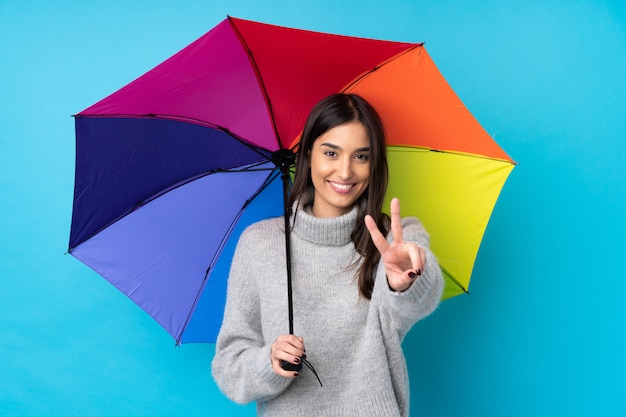 Young brunette woman holding an umbrella over blue wall smiling and showing victory sign