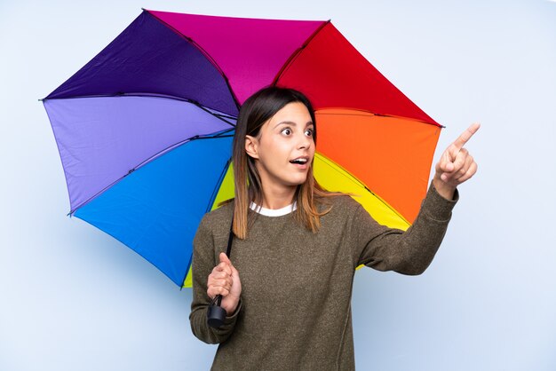 Young brunette woman holding an umbrella over isolated blue wall pointing away