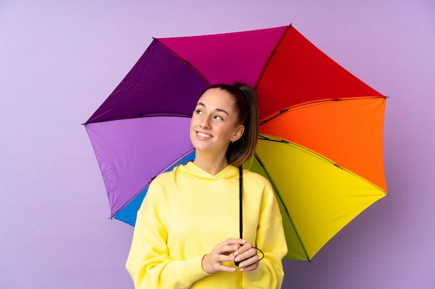 Young brunette woman holding an umbrella over isolated purple wall looking up while smiling