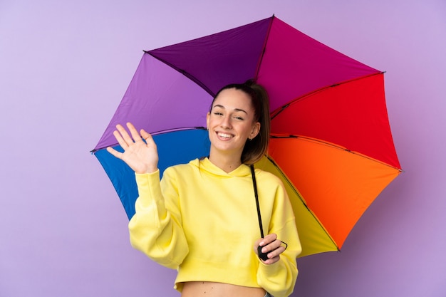 Young brunette woman holding an umbrella over isolated purple wall saluting with hand with happy expression