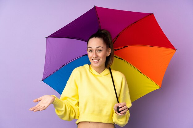 ショックを受けた表情で分離された紫色の壁に傘を置く若いブルネットの女性