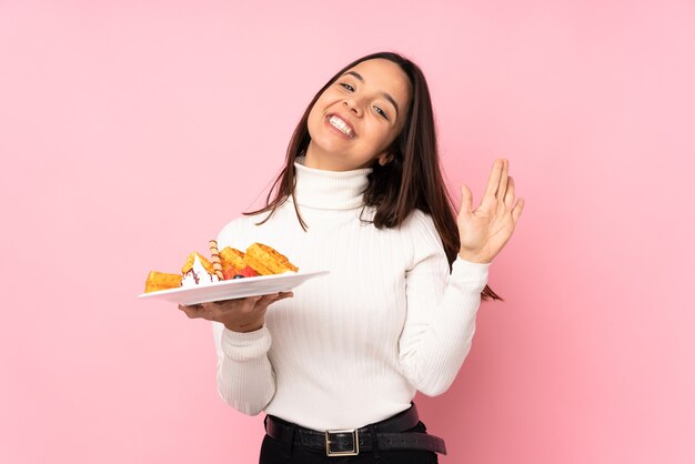 Young brunette woman holding waffles on isolated pink saluting with hand with happy expression