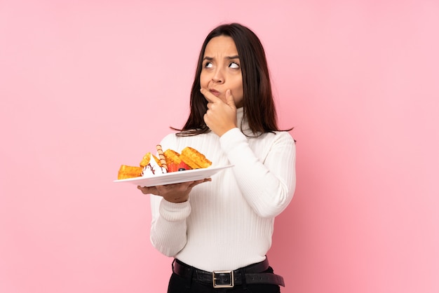 Young brunette woman holding waffles over isolated pink wall having doubts and with confuse face expression