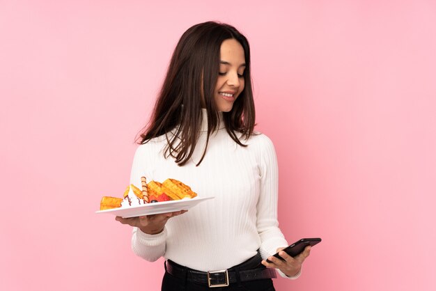 Young brunette woman holding waffles over isolated pink wall holding coffee to take away and a mobile