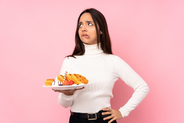 Young brunette woman holding waffles over isolated pink wall with confuse face expression