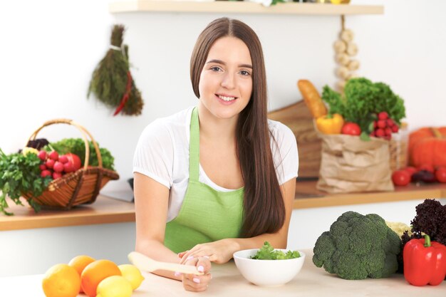 Young brunette woman is cooking or eating fresh salad in the kitchen. Housewife holding wooden spoon in her right hand. Food and health concept.