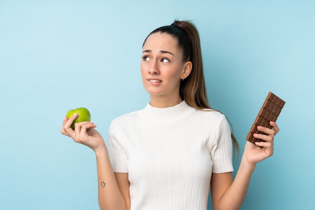 Молодая брюнетка женщина над изолированной синей стеной, имея сомнения, принимая таблетку шоколада в одной руке и яблоко в другой