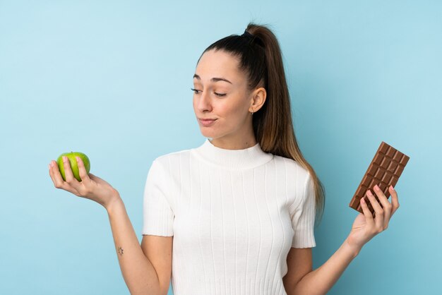 Young brunette woman over isolated blue wall having doubts while taking a chocolate tablet in one hand and an apple in the other