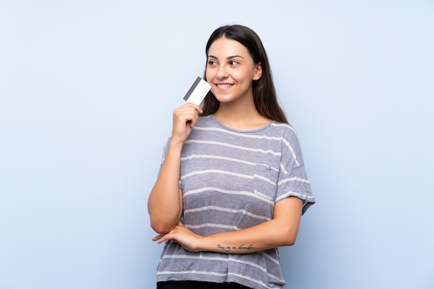 Young brunette woman over isolated blue wall holding a credit card