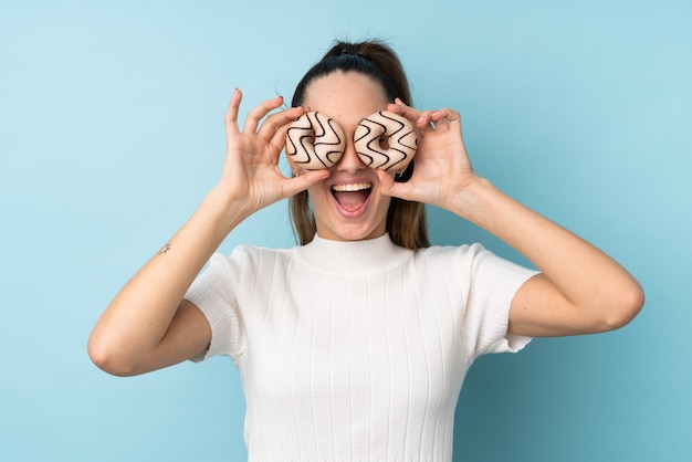 Young brunette woman over isolated blue wall holding donuts in an eye