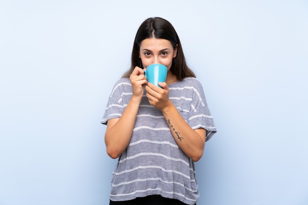 커피의 뜨거운 컵을 들고 고립 된 파란색 벽에 젊은 갈색 머리 여자