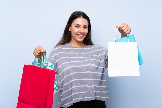 Young brunette woman over isolated blue wall holding a lot of shopping bags