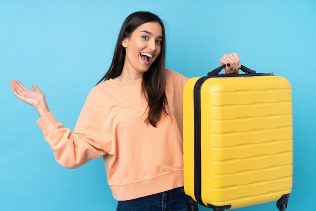 Young brunette woman over isolated blue wall in vacation with travel suitcase and surprised