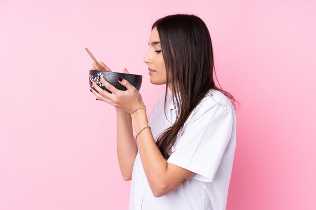 Young brunette woman over isolated pink wall holding a bowl of noodles with chopsticks and eating it