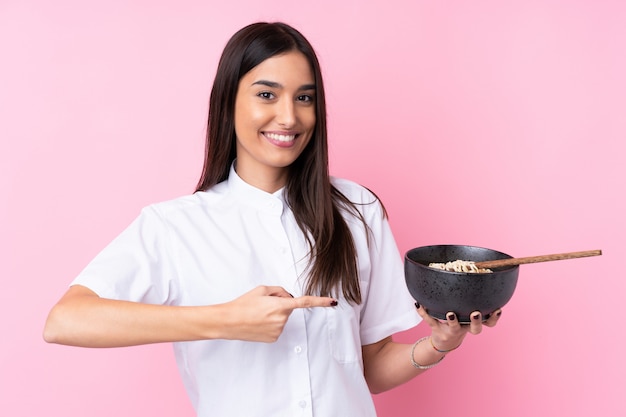 Giovane donna castana sopra la parete rosa isolata e indicarla mentre tiene una ciotola di tagliatelle con le bacchette