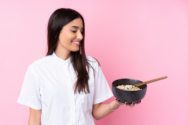 Young brunette woman over isolated pink wall with happy expression while holding a bowl of noodles with chopsticks