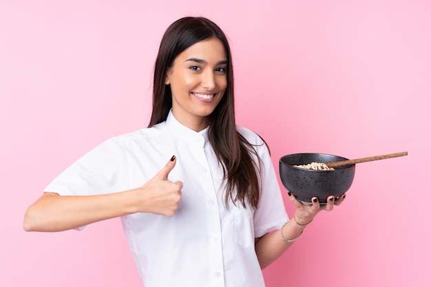 箸で麺のボウルを保持しながら何か良いことが起こったので、親指で孤立したピンクの壁の上の若いブルネットの女性