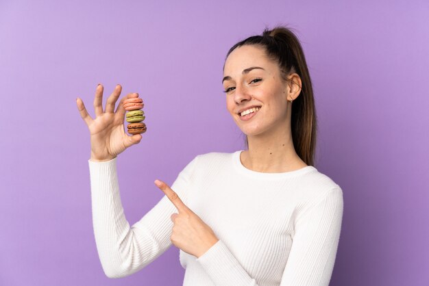 Young brunette woman over isolated purple wall holding colorful french macarons and pointing it