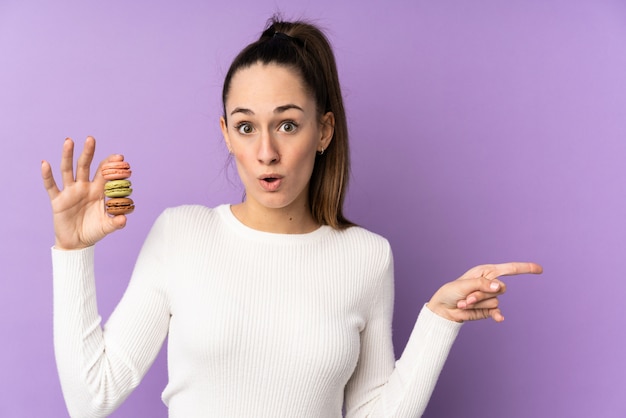 Giovane donna castana sopra la parete porpora isolata che tiene i macarons francesi variopinti e che indica lato