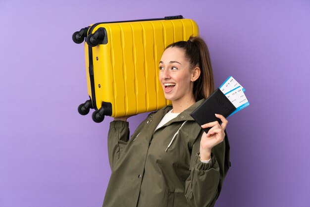 Young brunette woman over isolated purple wall in vacation with suitcase and passport