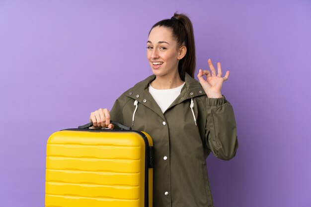 Young brunette woman over isolated purple wall in vacation with travel suitcase and a hat