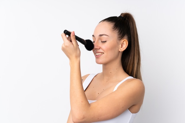 Young brunette woman over isolated white wall holding makeup brush and whit happy expression