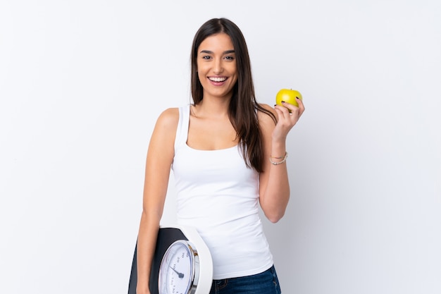 Young brunette woman over isolated white wall with weighing machine and with an apple