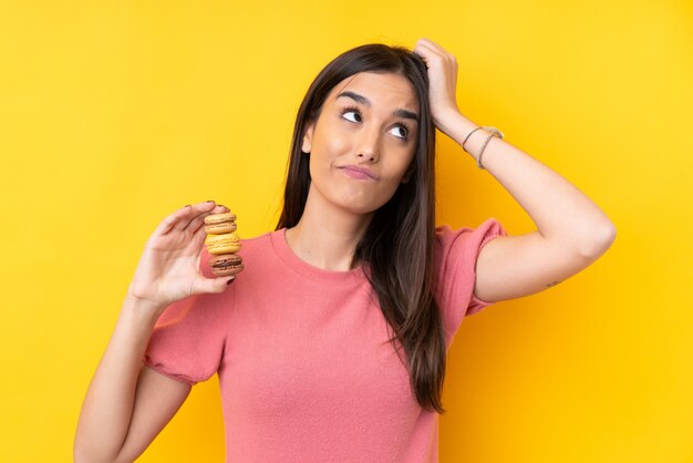 Young brunette woman over isolated yellow wall holding colorful French macarons with confuse expression