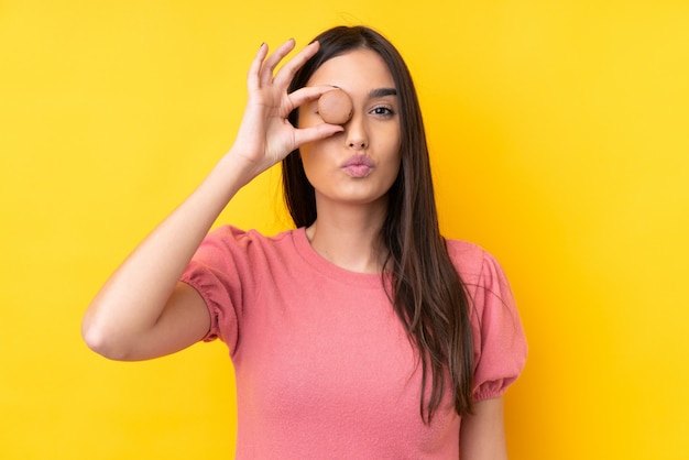 Young brunette woman over isolated yellow wall holding colorful French macarons with funny face