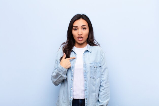 young brunette woman looking shocked and surprised with mouth wide open pointing to self