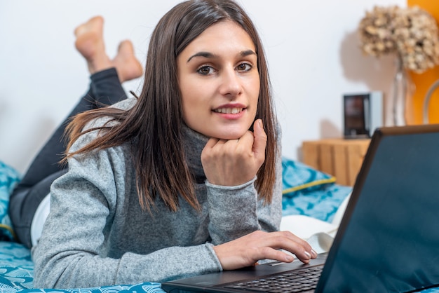 젊은 갈색 머리 여자는 노트북을 사용하여 침대에 누워