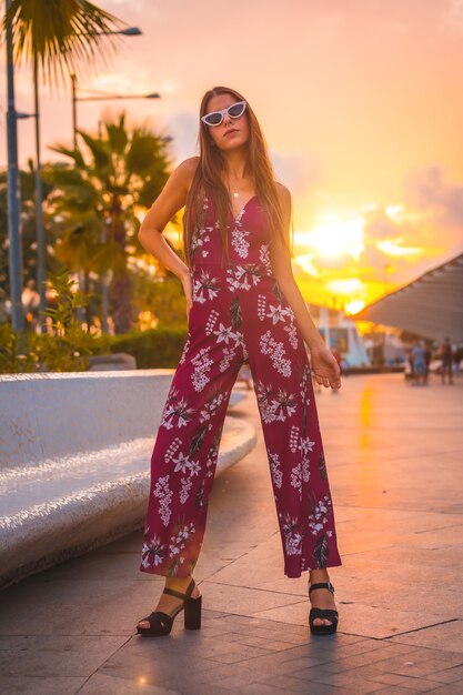 Young brunette woman in a maroon floral dress and modern sunglasses enjoying the summer in the golden hour