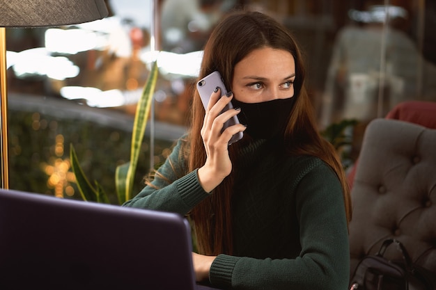 Giovane donna castana in mascherina medica nella caffetteria con laptop e smartphone