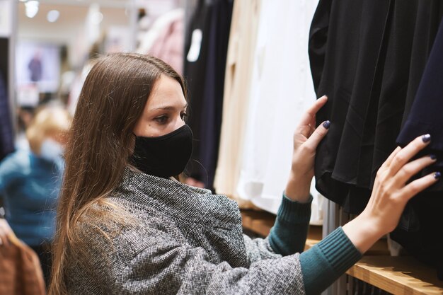 Young brunette woman in medical mask chooses clothes in the store and makes purchases