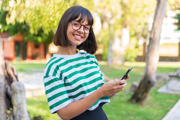 Молодая брюнетка в парке отправляет сообщение или электронное письмо с мобильного телефона