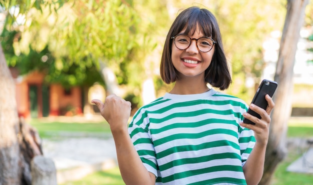 Young brunette woman in the park using mobile phone and pointing to the lateral