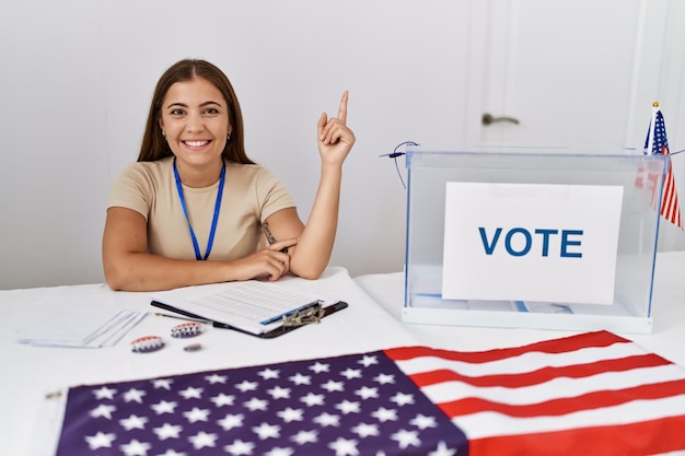 Young brunette woman at political election sitting by ballot smiling happy pointing with hand and finger to the side