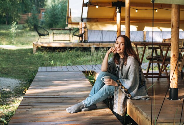 Photo young brunette woman in poncho drinking tea and relaxing in glamping in nature