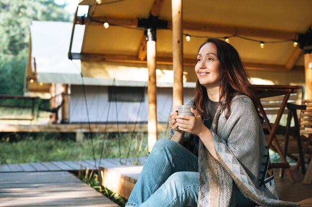 ポンチョでお茶を飲み、自然の中でグランピングでリラックスした若いブルネットの女性