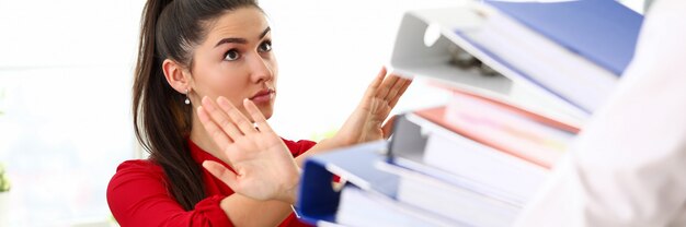 Young brunette woman in red shirt sitting in office and refusing to work with heap of documents.